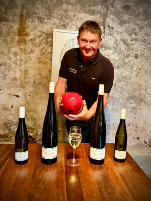 John Hughes holding footy in front of bottles of Riesling on a wooden table